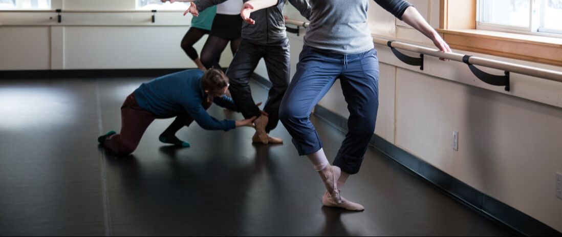 Four adult ballet dancers are visible from the waist down. They are wearing exercise clothes and t-shirts and stand facing the camera with their left hands on the barre, their left legs bent and their right feet pointed at their left ankles. The instructor squats down to adjust the right foot of the second dancer in line using both hands to shape the foot.