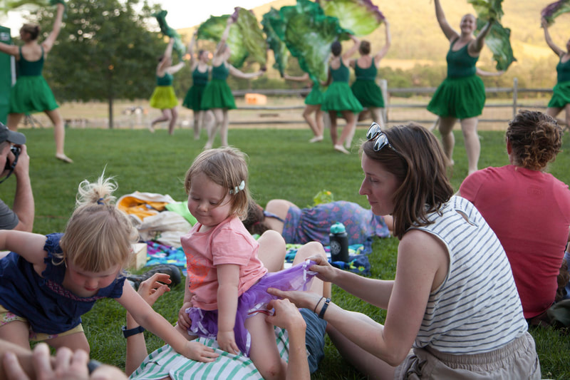In the foreground, parents and their excited kids in a tutu and summer-wear are sitting in the grass in the audience of a Farm to Ballet Performance as dancers in green leotards and skirts holding huge fake lettuce leaves overhead make a dancing salad.