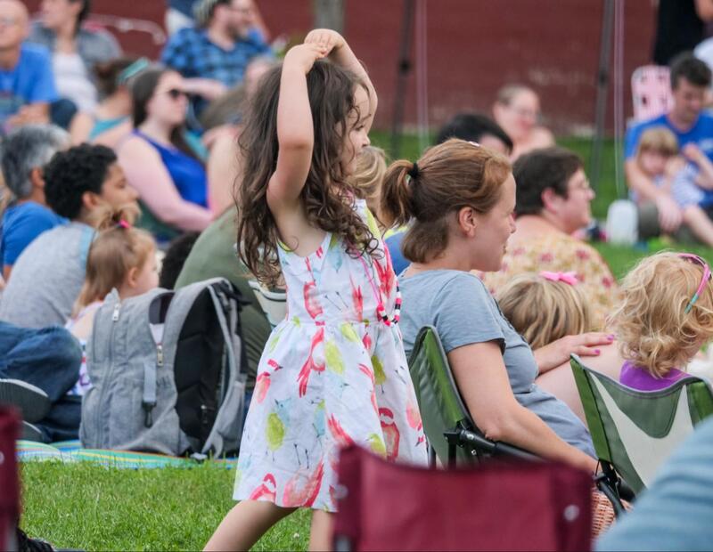 A child with long brown hair and a floral dress raises both hands around her head and dances, imitating a ballerina as she watches the show. Around her other audience members sit on blankets and lawn chairs looking transfixed by the performance.