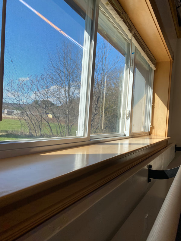A view out the window by the barre at Ballet Vermont. There is a bright blue sky with a couple fluffy white clouds and green grass visible behind a tall budding bush. The window ledge is wide and light wood and there are shadows cast at a diagonal to the right.