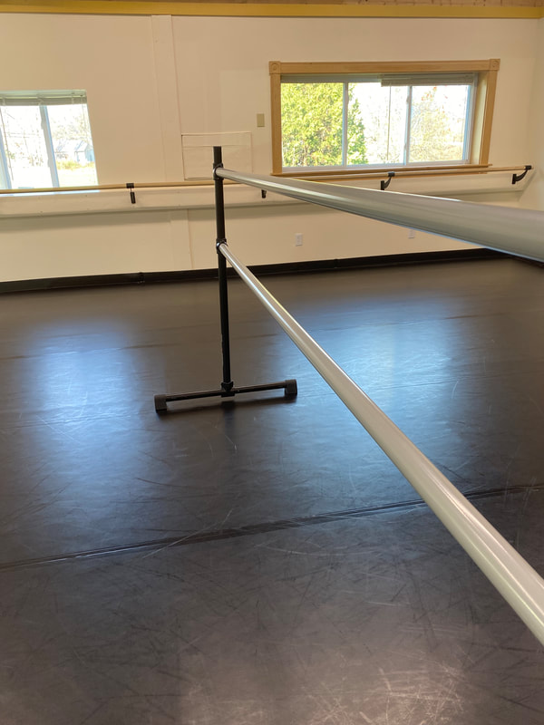 A close view along a freestanding ballet barre. The barre extends from the middle of the photo out through the right foreground of the frame. Behind the barre there are two windows with bright sunshine reflecting up off the marley floor. 