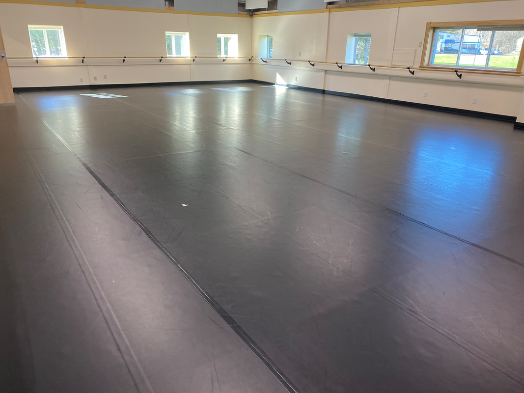 This picture shows almost the entire studio room of Ballet Vermont. There is a big black marley floor and a barre that runs along the two walls shown. Bright Vermont sunshine streams through the six windows above the barre. Visible through the windows are trees and a green lawn.