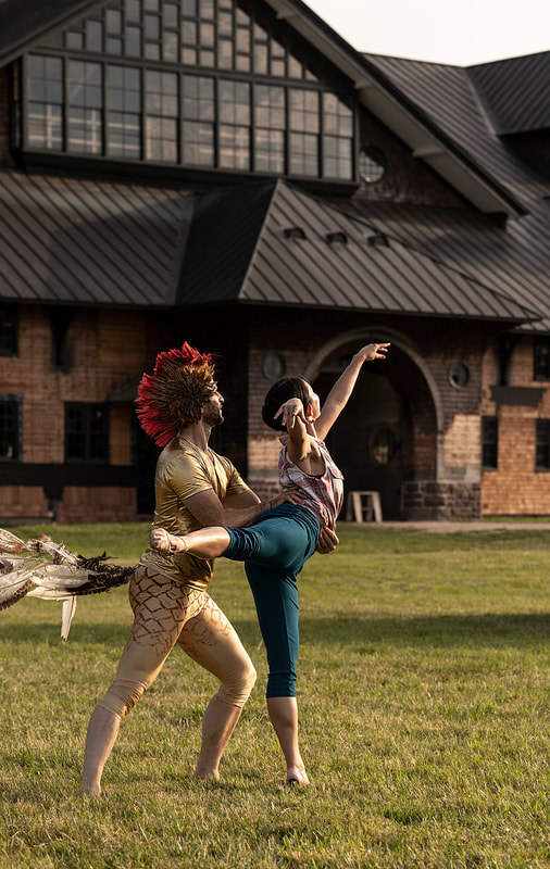 A dancer dressed as a rooster in a golden shirt and pants, feather tail, and red mohawk headpiece holds the waist of a dancer dressed as a farmer in bright blue pants and gingham tank top as she lifts he leg and arm to create a beautiful arabesque. They are in front of an elegant wooden barn and looking into the distance. 