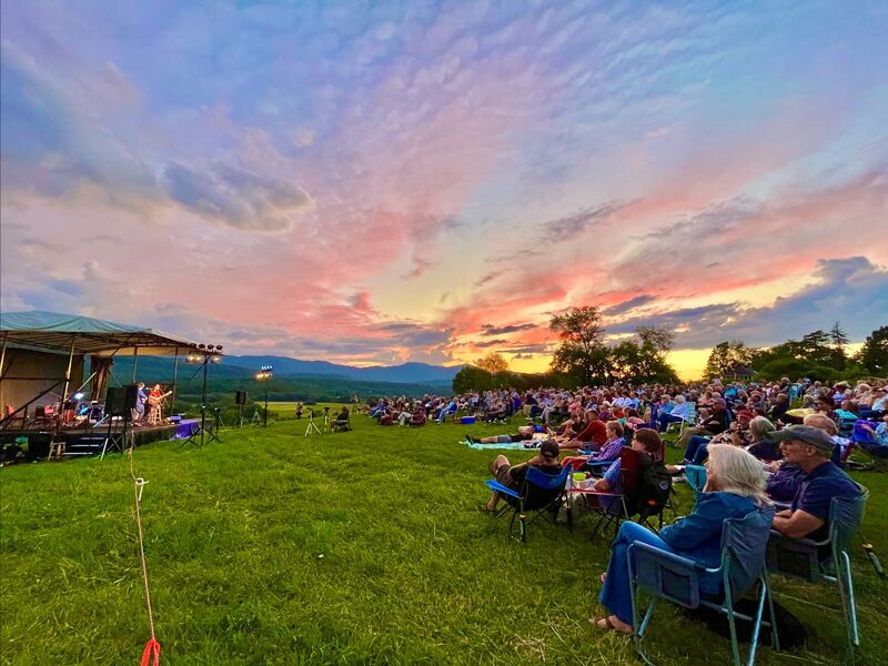 A large audience sits in lawn chairs on the expansive green grass. The crowd is on the right side facing an outdoor stage on the left with a performer on it. The sunset sky is beautiful with streaks of pink, purple, blue, and yellow, and it takes up half of the image.